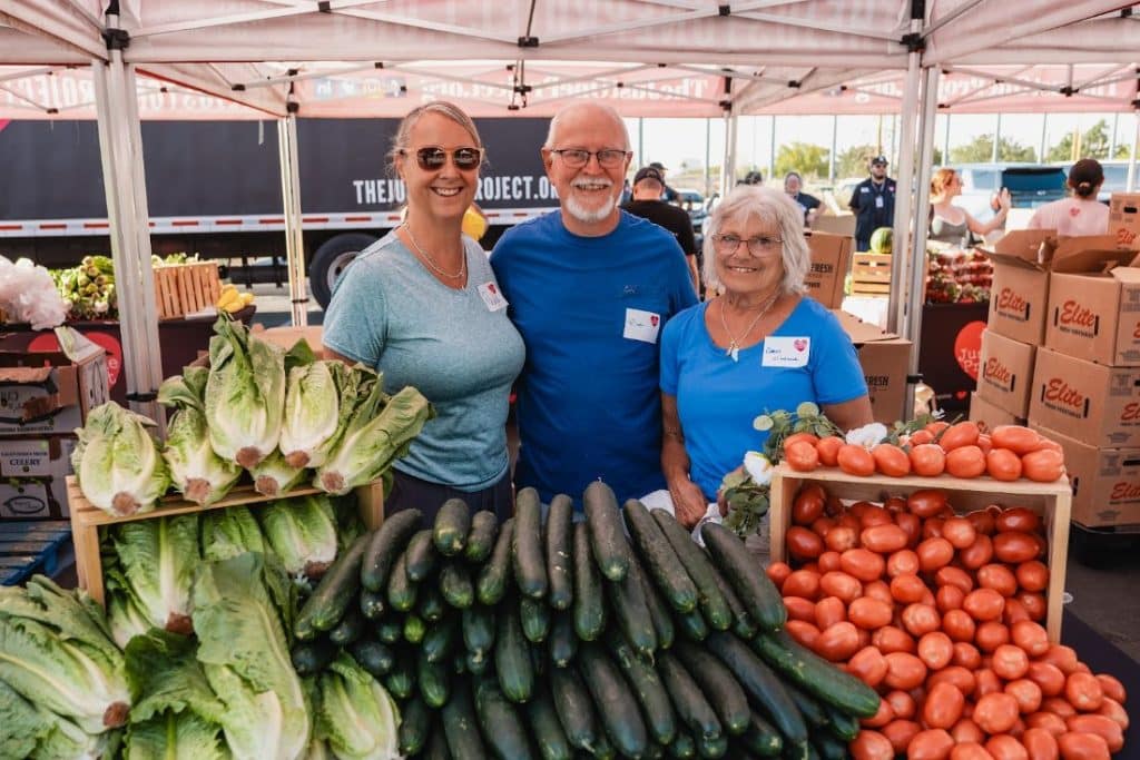 Three smiling people stand behind a table laden with fresh lettuce, cucumbers, and tomatoes at an outdoor market.