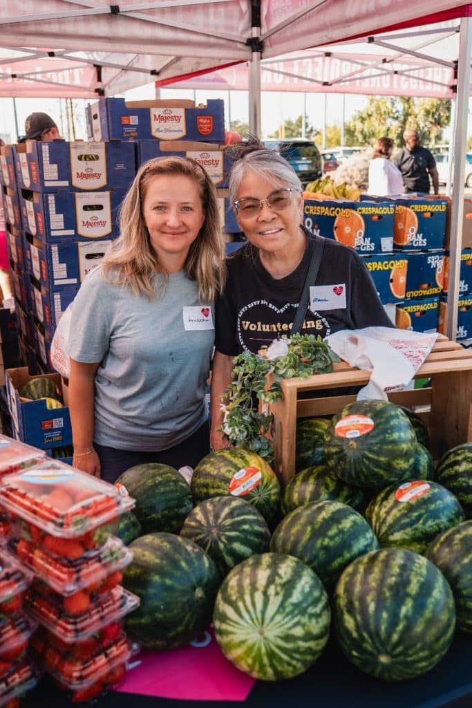 Two smiling women stand behind a table laden with watermelons and strawberries at an outdoor market.