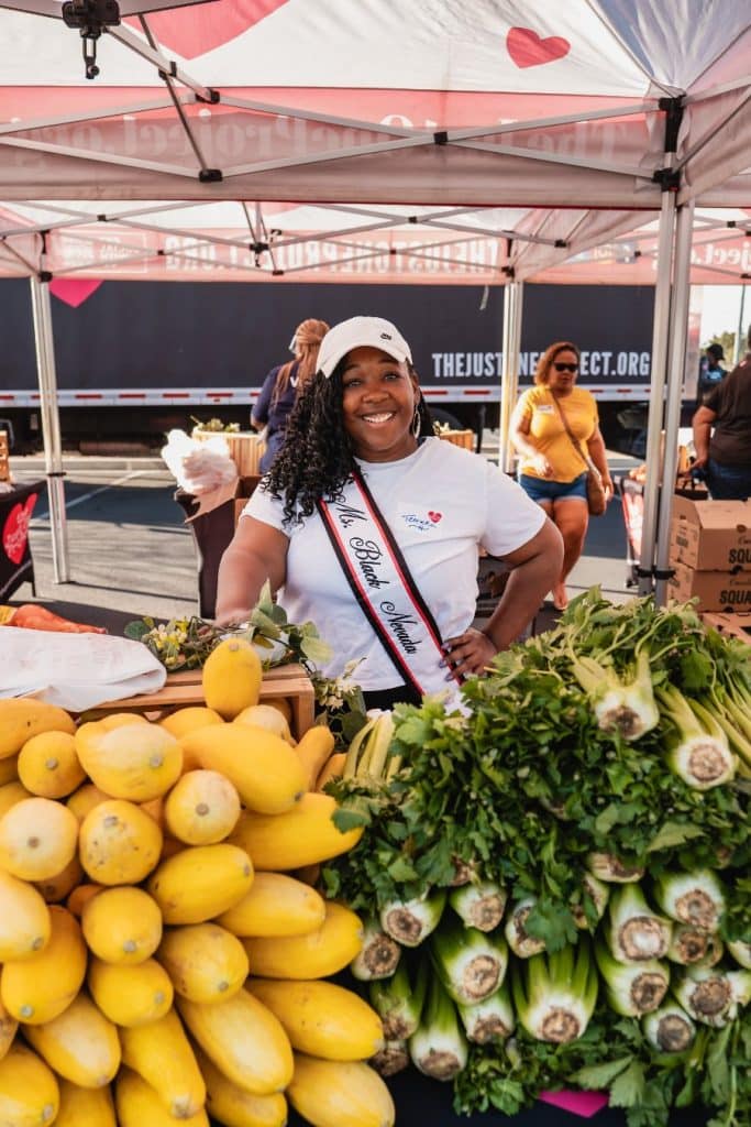 A smiling woman wearing a "Ms. Black Nevada" sash stands behind a table of yellow squash and green celery.