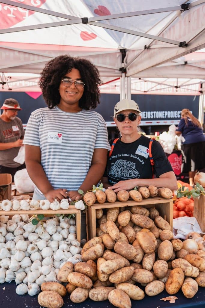 Two women stand behind tables laden with garlic and potatoes at an outdoor market.