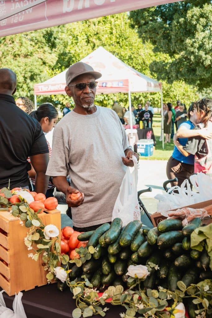 A man in a gray shirt and sunglasses holds a tomato and a plastic bag at a produce stand.