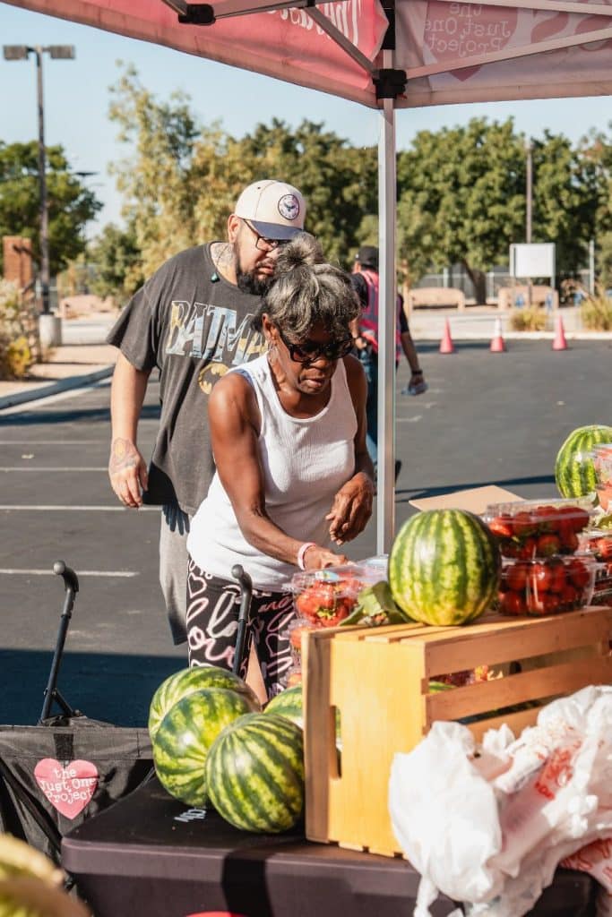 A woman in sunglasses and a man in a "BATMAN" shirt stand behind a table with watermelons and strawberries.