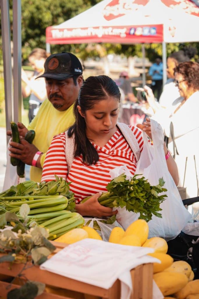 A young woman in a red and white striped shirt holds a bunch of parsley at a produce stand.