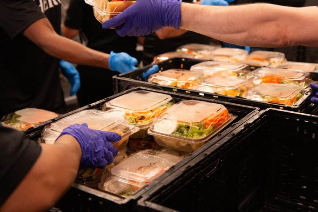 Several people wearing blue and purple gloves are packing clear plastic containers of food into black crates.