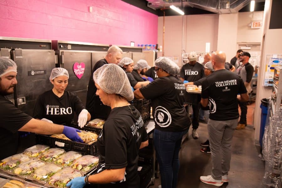 Volunteers in hairnets and black "HERO" shirts prepare packaged meals in a bustling kitchen.