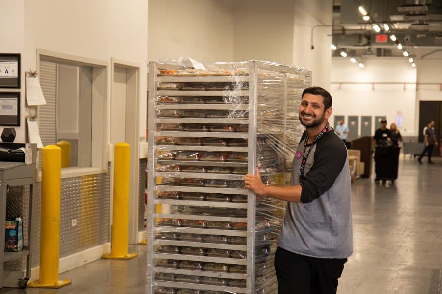 A smiling man in a gray and black shirt pushes a tall, metal shelving unit full of wrapped food containers.