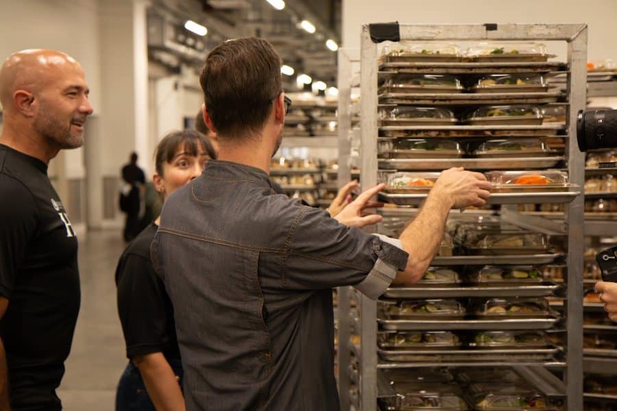 A man in a chef's jacket places a tray of packaged meals onto a tall metal rack, while two people watch.