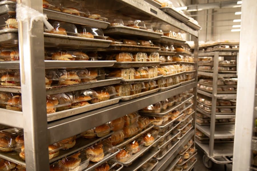 Multiple metal racks are filled with rows of individually packaged sandwiches and pastries in a commercial kitchen.