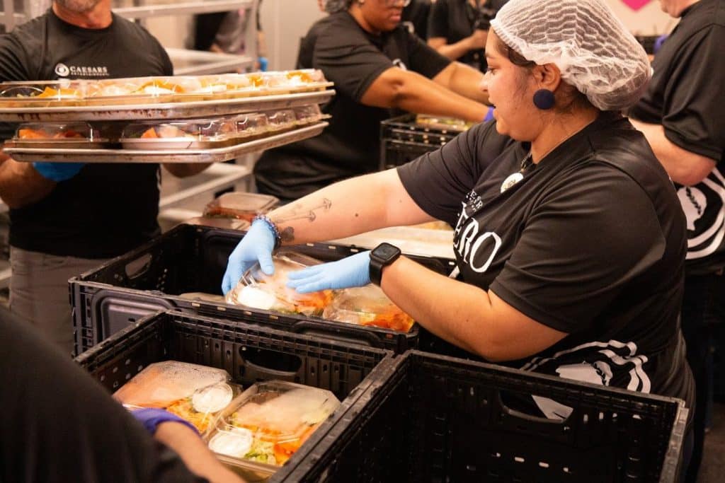 People in black shirts and blue gloves pack clear containers of food into black crates.