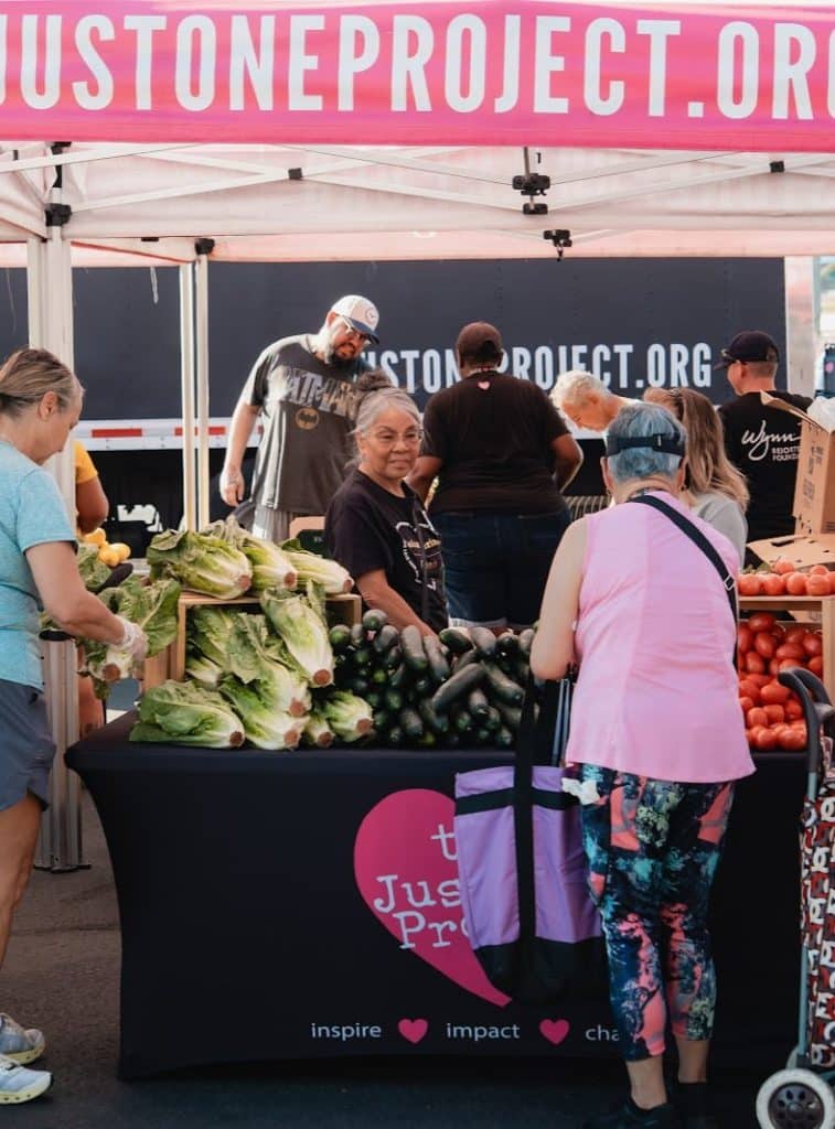 A woman sits behind a table piled with lettuce and cucumbers, with a "JUSTONEPROJECT.ORG" banner above.