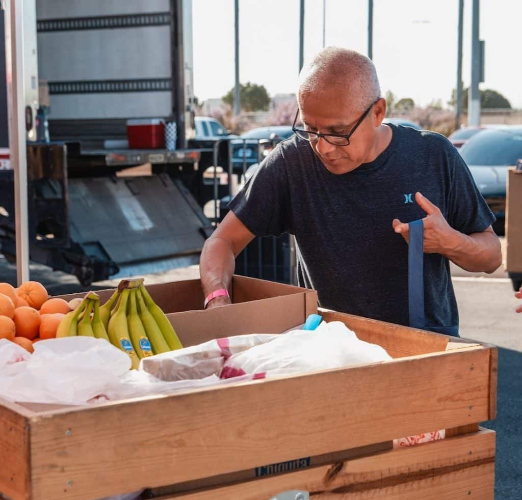 A man in glasses and a dark gray "Hurley" t-shirt sorts items in a wooden crate of fruit.