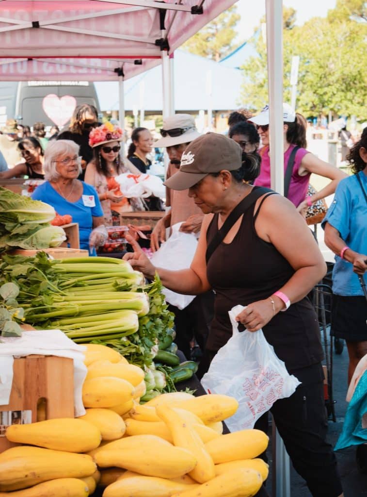 A woman in a brown cap and tank top shops for celery at a bustling farmers market.