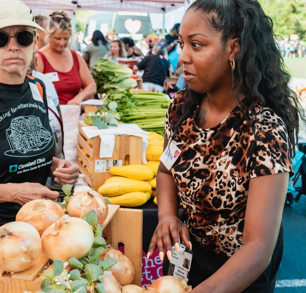 A woman in a leopard print shirt stands at a produce stall with onions, squash, and greens.