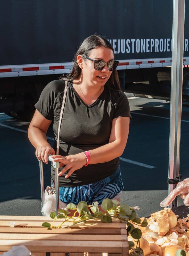 A woman in sunglasses and a black shirt stands by a market stall with onions, "EJUSTONEPROJE" on a truck behind her.