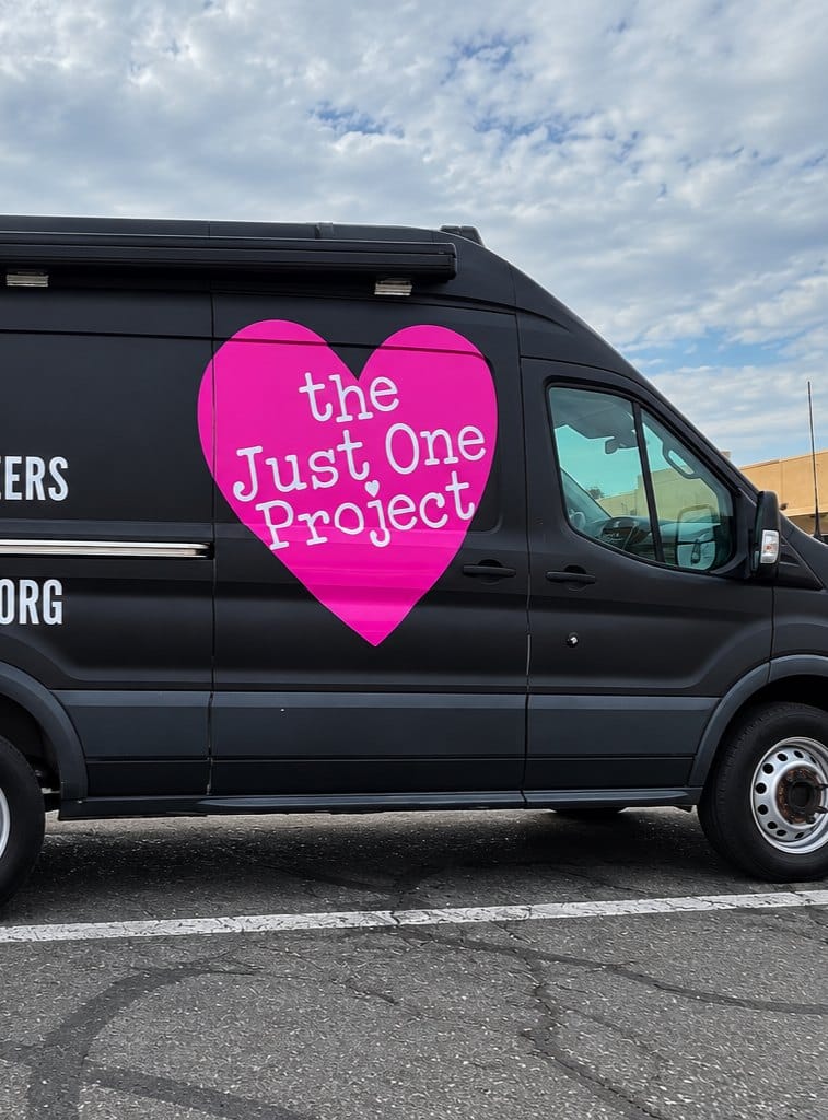 A black van with a large pink heart logo reading "the Just One Project" on its side, parked on asphalt.