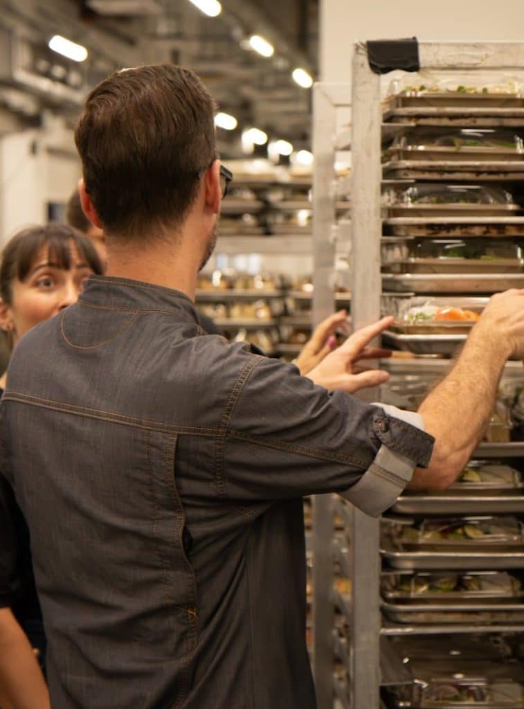 A man in a denim shirt reaches for a meal tray from a metal rack, with a woman looking on.