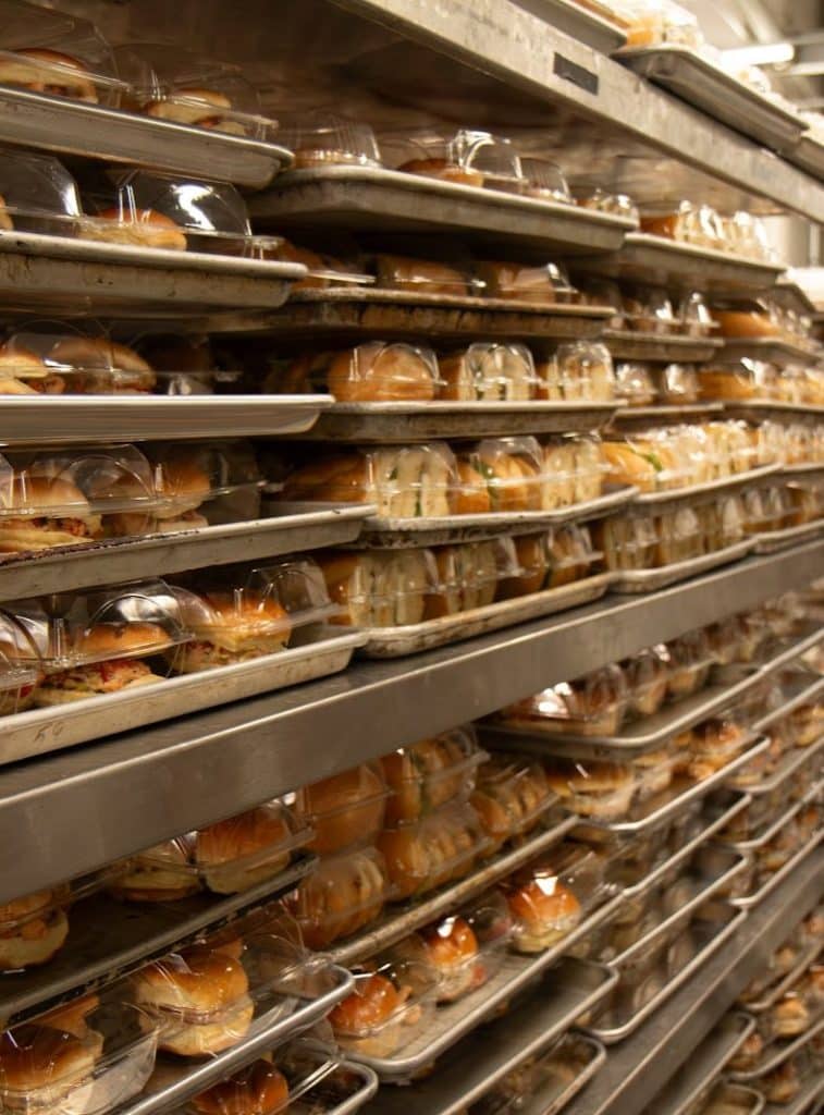 Numerous trays of packaged sandwiches and pastries are stacked on metal shelves in a commercial kitchen.