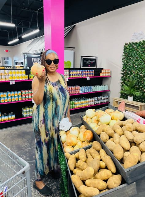 A smiling woman in sunglasses and a tie-dye dress holds a potato while standing by bins of produce in a grocery store.