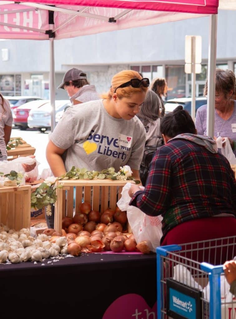 A woman in a gray "Serve Liberty" t-shirt helps a customer bag onions and garlic at an outdoor market.