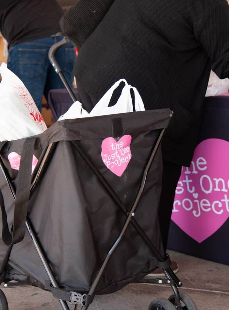 A black collapsible cart with white bags and a pink heart sticker reading "the Just One Project" is next to a person.