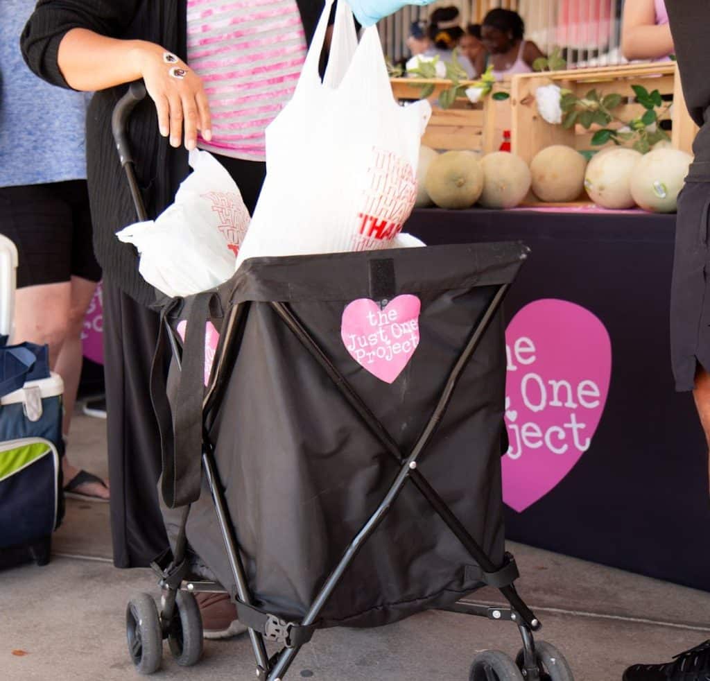 A person pushes a black shopping cart with white bags and a pink heart logo that reads "the Just One Project."