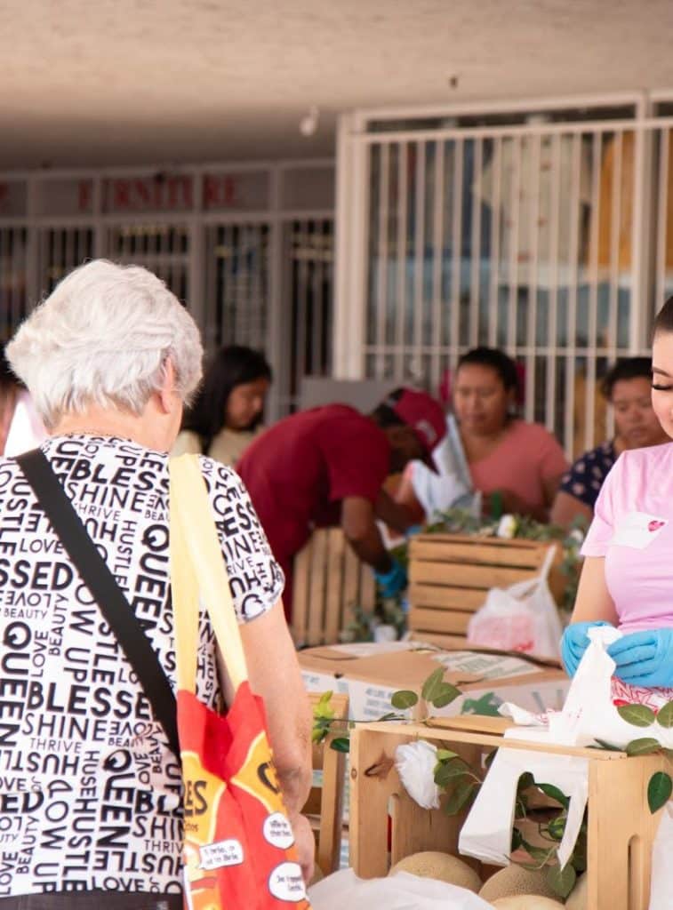 A person with white hair and a "BLESS SHINE THRIVE HUSTLE QUEEN" shirt looks at a market stall.