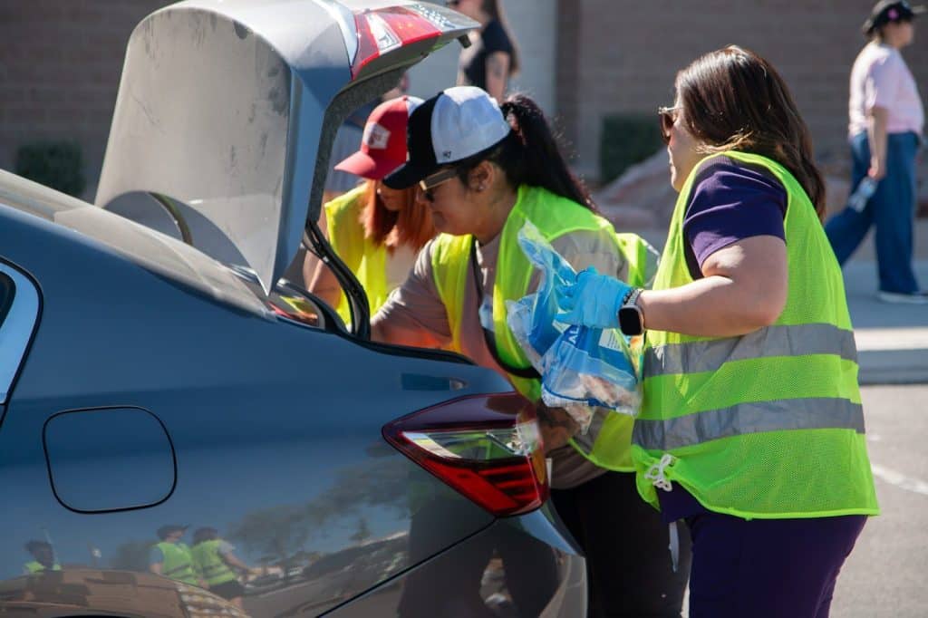 Three women in neon yellow safety vests load items into the open trunk of a gray car.