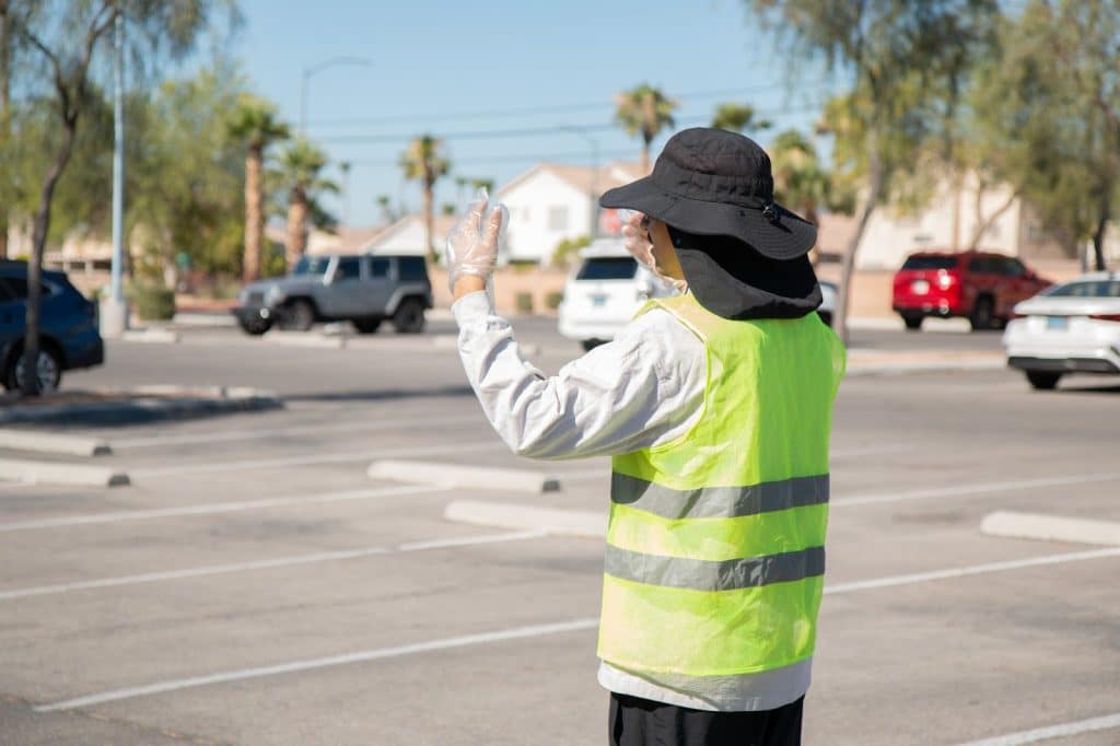A person in a black sun hat and reflective yellow vest directs traffic in a sunny parking lot.