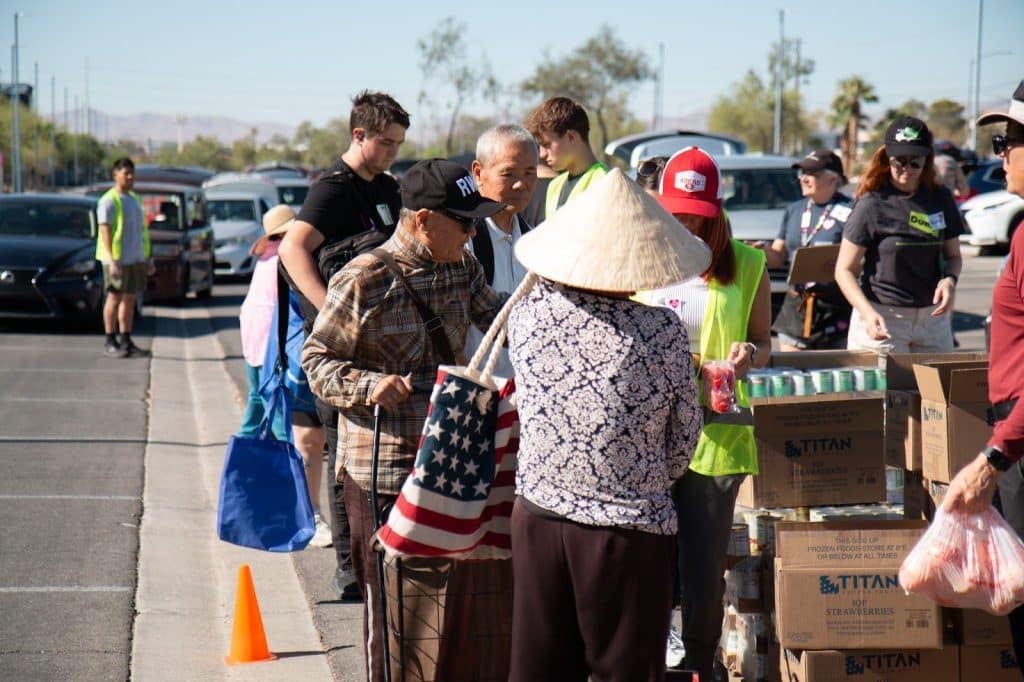 People line up outdoors for food distribution, with volunteers and boxes of "TITAN" brand goods.