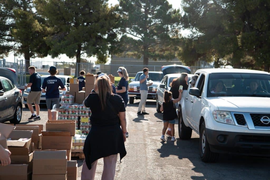 Volunteers, some wearing "SERVICE CREW" shirts, distribute food from boxes and pallets to people in cars.