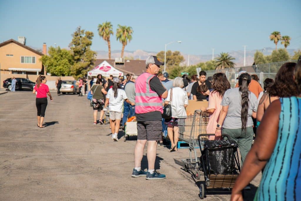 A man in a pink vest stands in a line of people with shopping carts in a sunny parking lot.