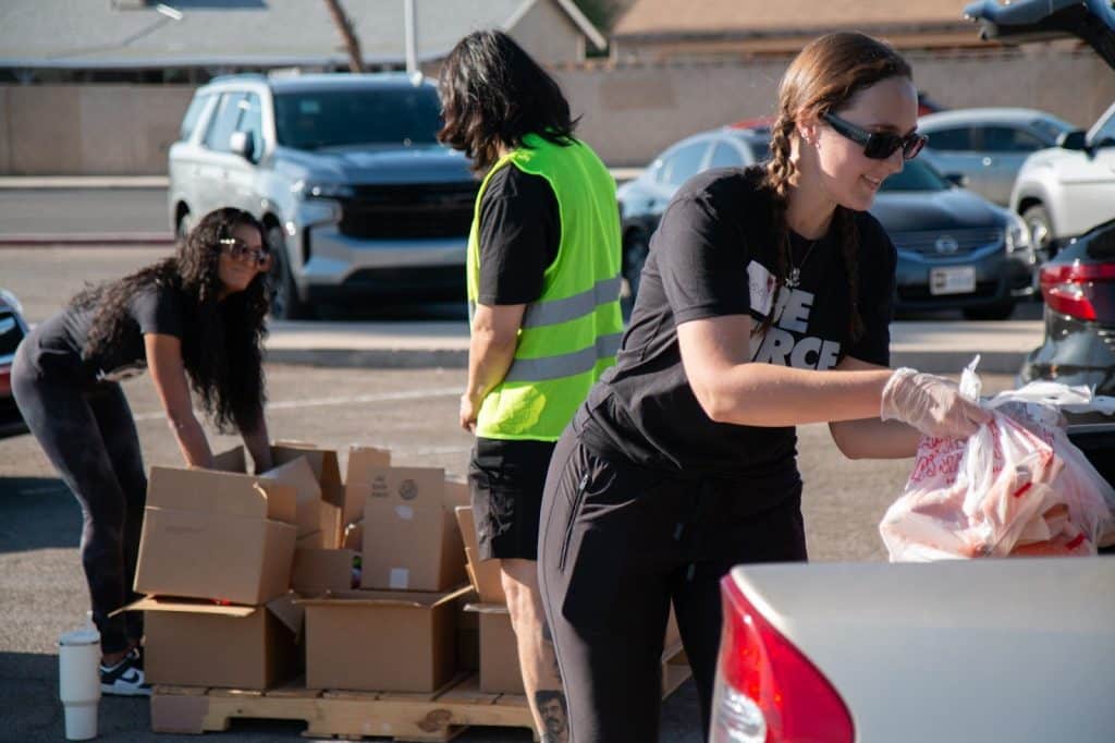 Three people, two women and one man, handle cardboard boxes and bags of goods in a parking lot.